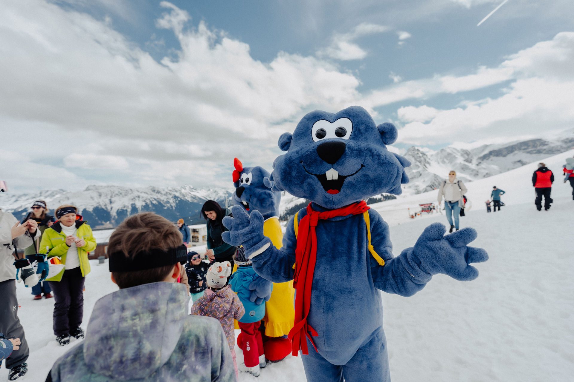 Two blue mascots with red scarves on a snowy mountain with children and adults, and snowy peaks in the background.
