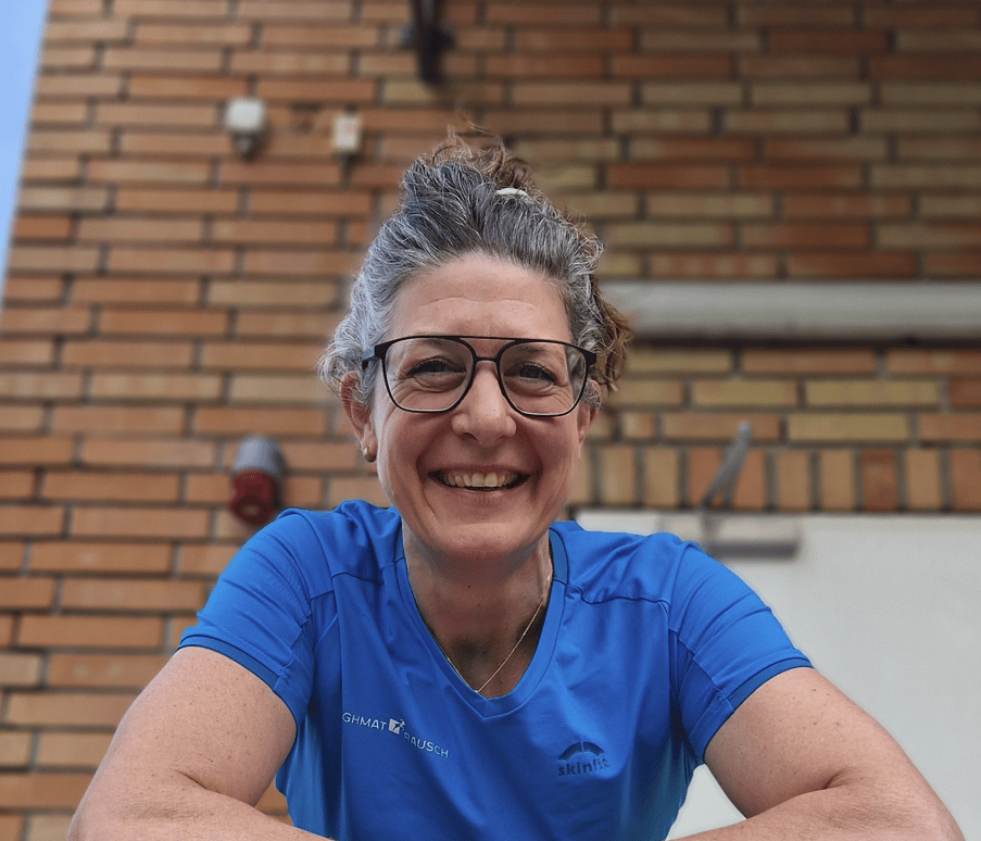 A woman with graying hair and glasses smiles brightly, wearing a blue t-shirt, against a brick wall.