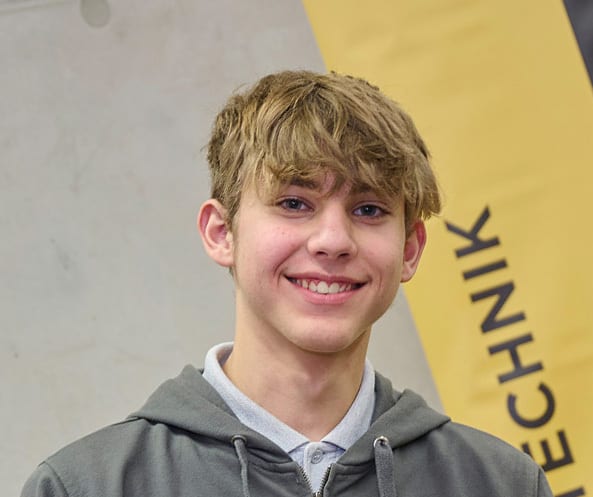 Smiling young man with light hair, wearing a grey hoodie. Yellow banner in background reads 'TECHNIK'.