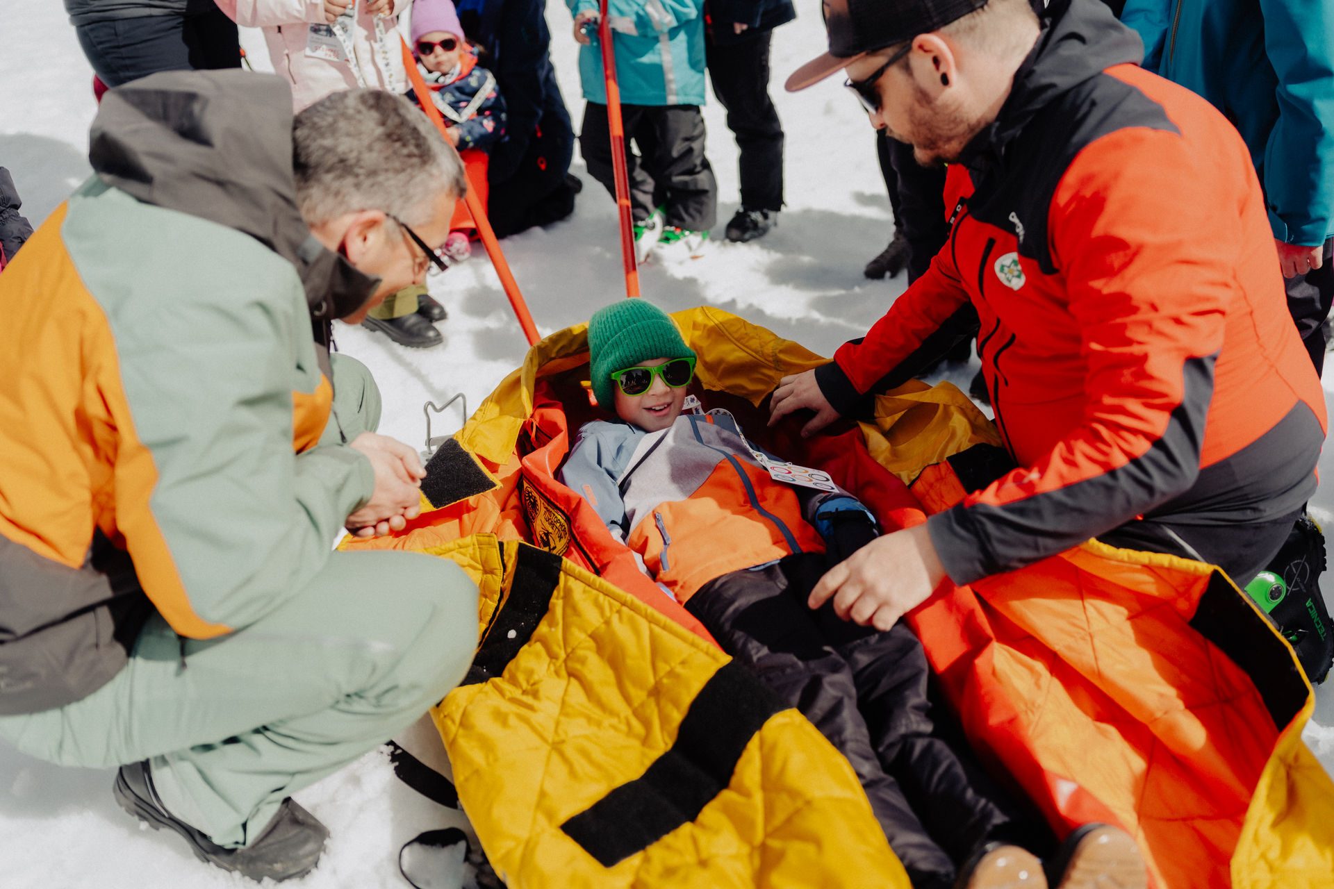 Child in a yellow sled on snow, tended by two adults in winter gear, smiling during a drill.