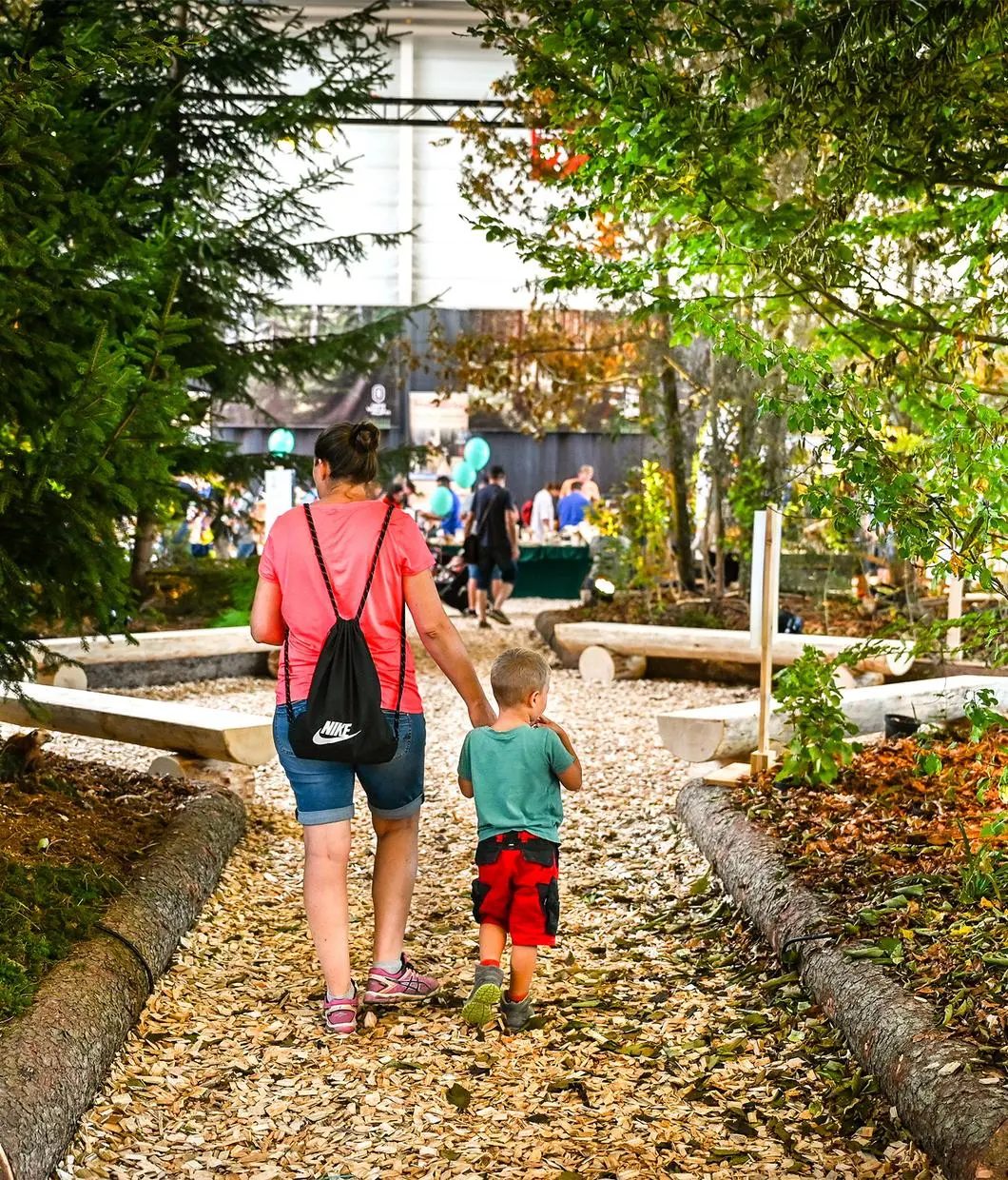 Mother and child walking hand-in-hand on a woodchip path through a park-like exhibition area.