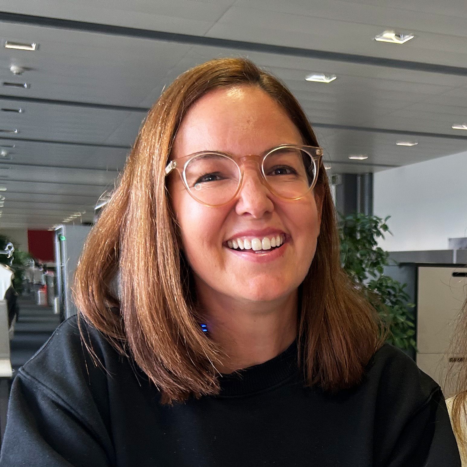 A smiling woman with brown hair and clear glasses, wearing a black top, in an office.