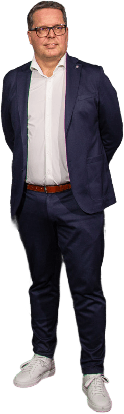 A man in glasses, navy suit, white shirt, brown belt, and white sneakers stands.