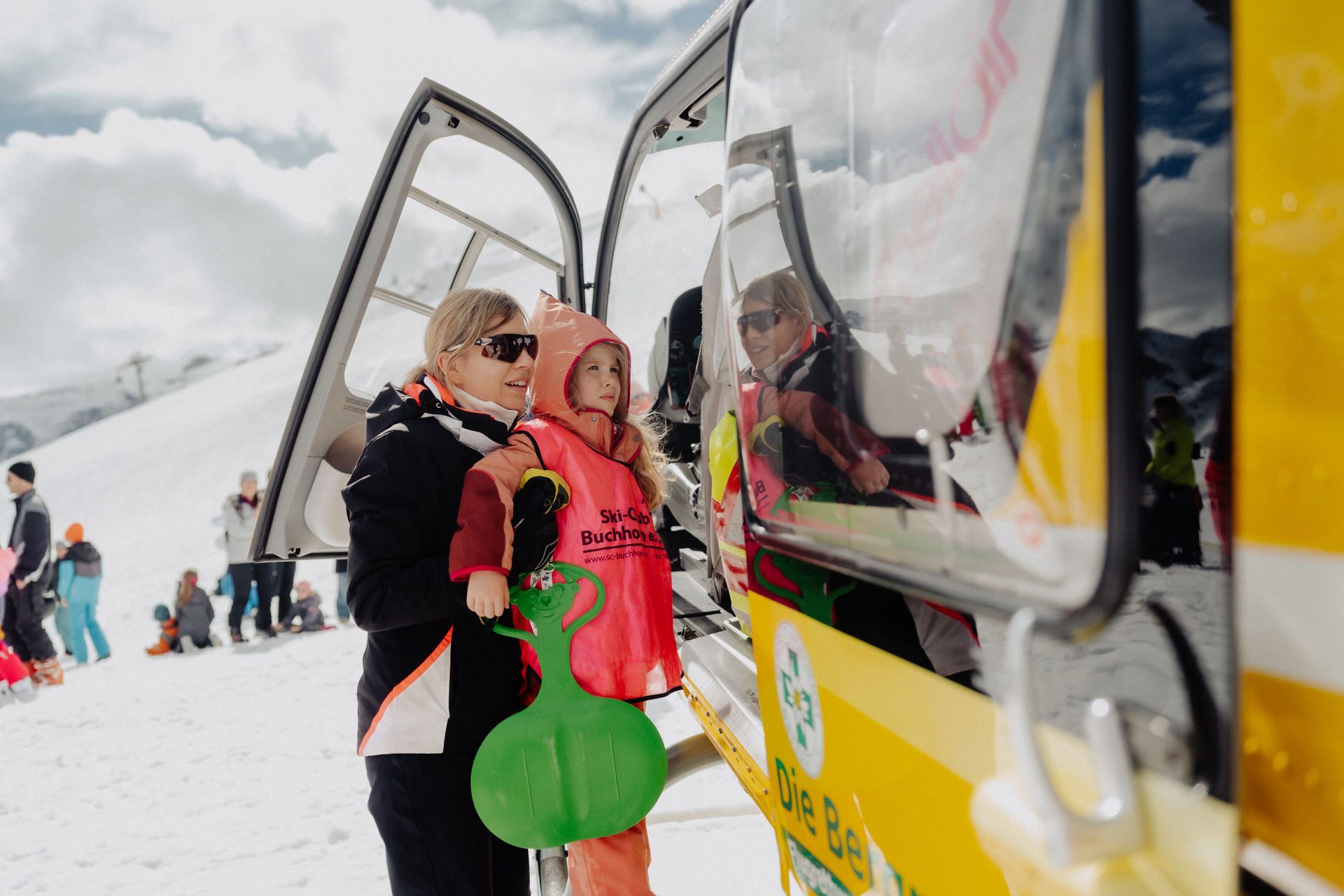 Woman and girl with a green sled by an open door of a yellow vehicle on a snowy mountain.