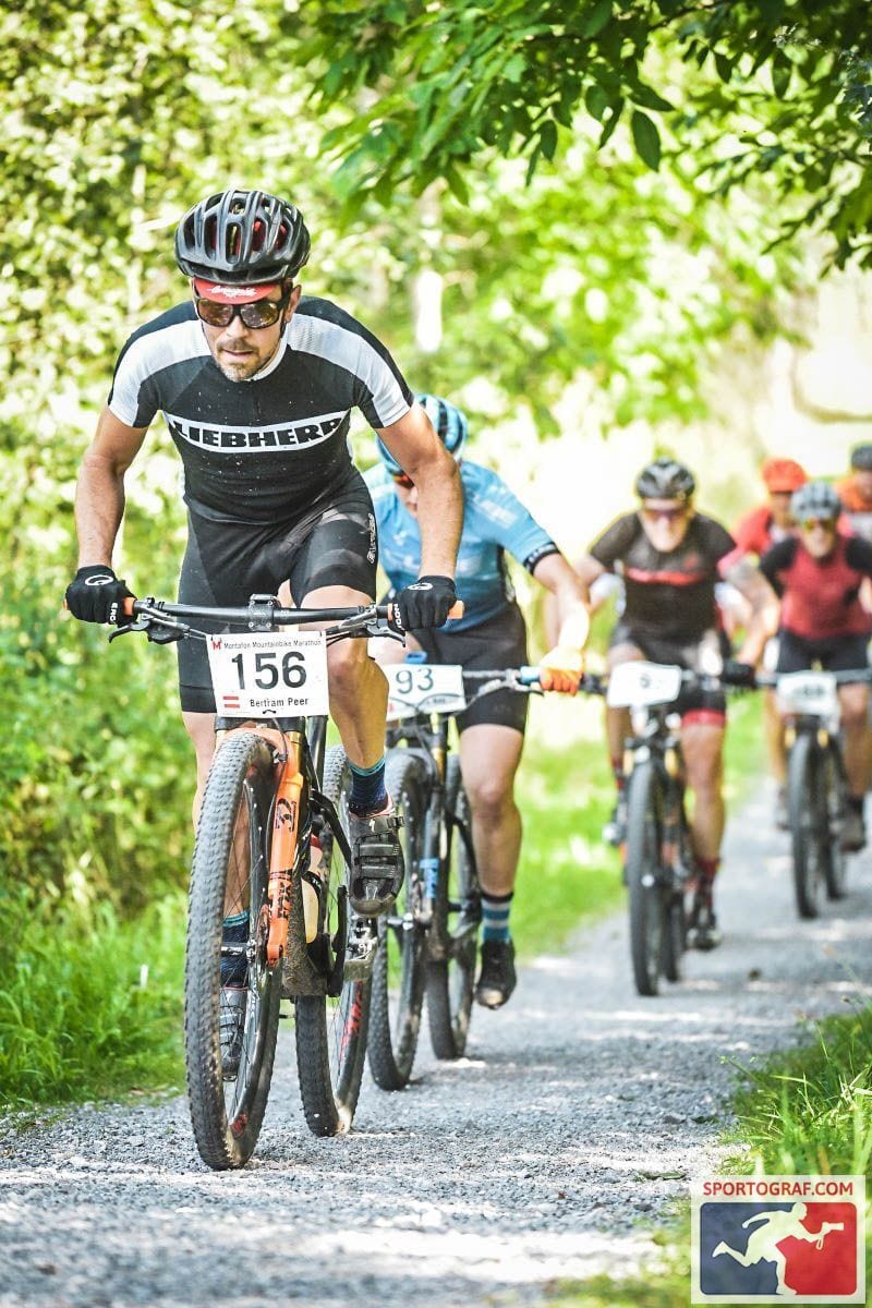 Mountain bikers race on a gravel path under green trees, led by a rider in a black and white jersey.