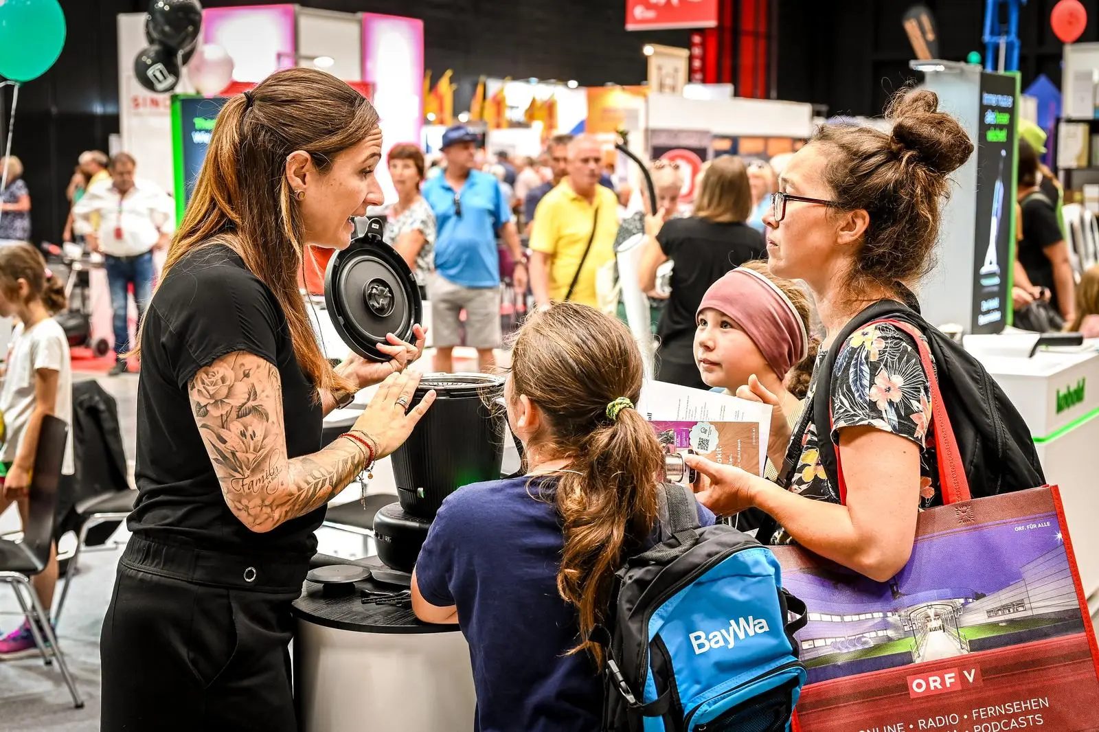 Woman demonstrates product to a mother and two children at a busy indoor event.