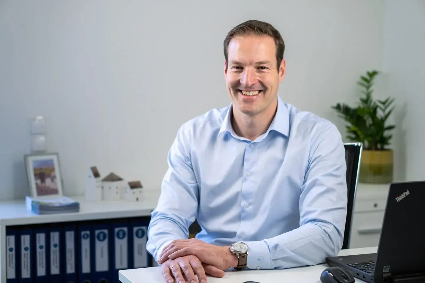 Smiling businessman in a light blue shirt sitting at a desk with a laptop and files in an office.