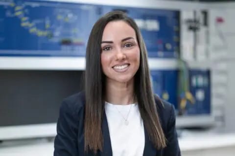 Smiling woman with dark hair wearing a jacket, against a blue technical panel.