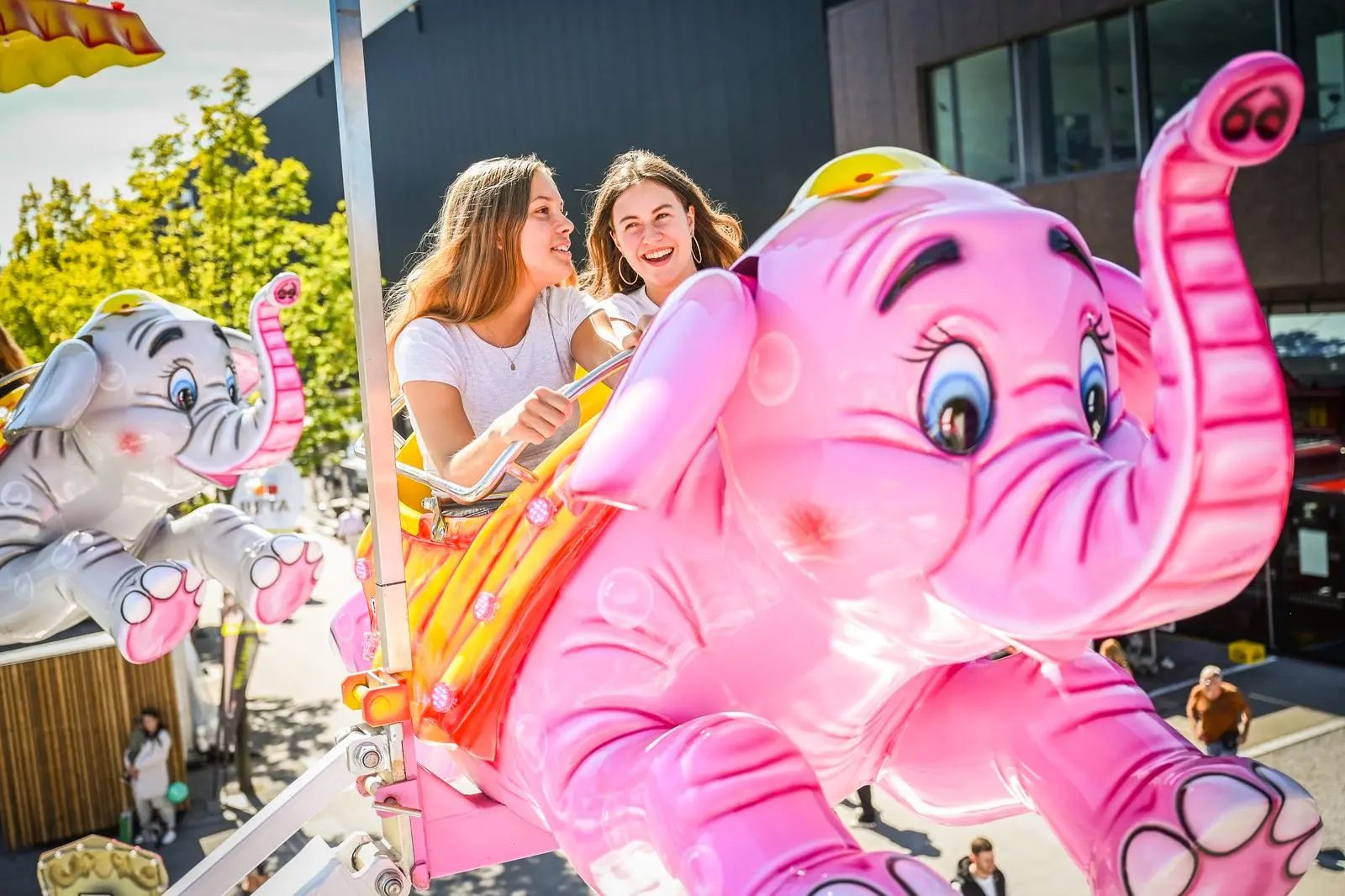 Two smiling young women enjoying a carousel ride featuring pink and grey elephant characters at a fair.