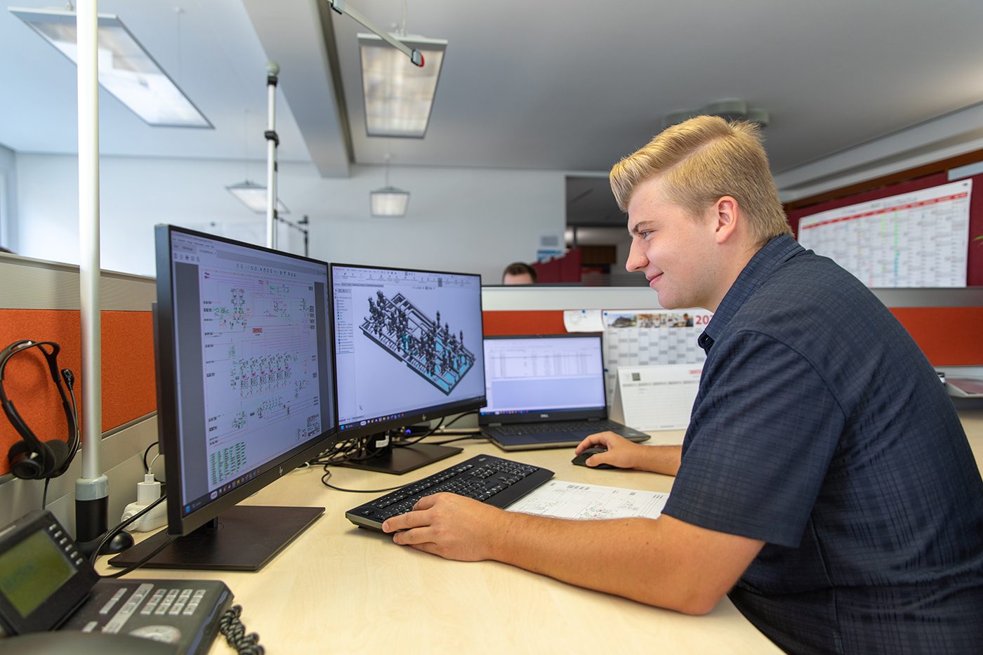 Smiling young man works at dual monitors showing technical designs.