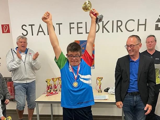 Young man with Down syndrome raises a gold trophy in triumph, surrounded by applauding adults.