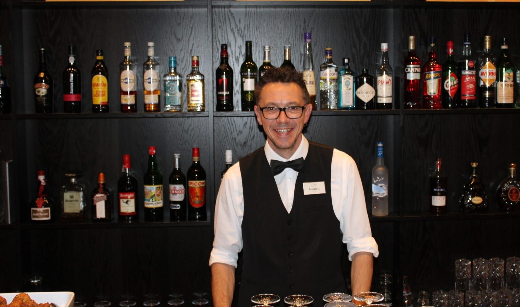 Smiling bartender Norbert behind a bar stocked with liquor bottles.