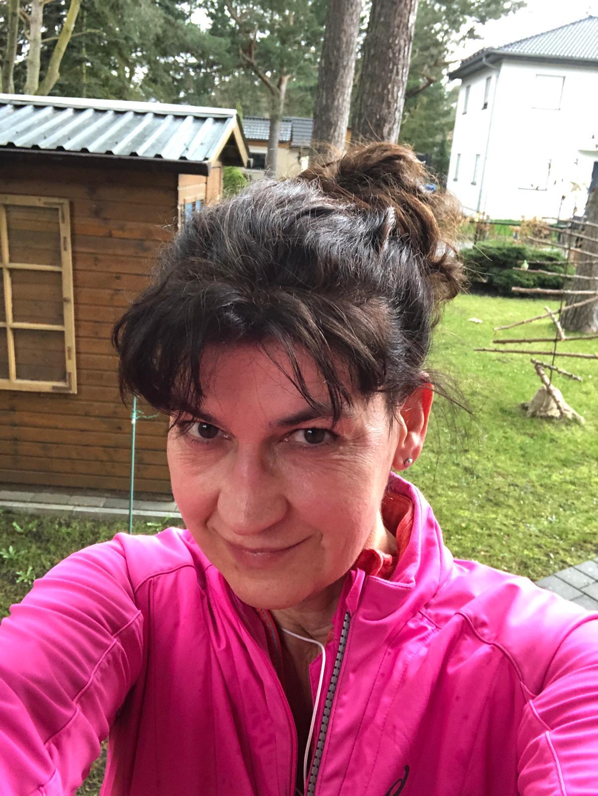 Smiling woman in a pink jacket with a messy bun, outdoors with houses and trees.