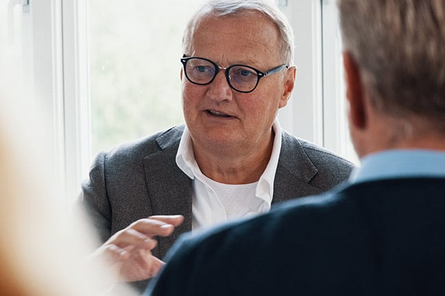 Older man in glasses speaks to another man.