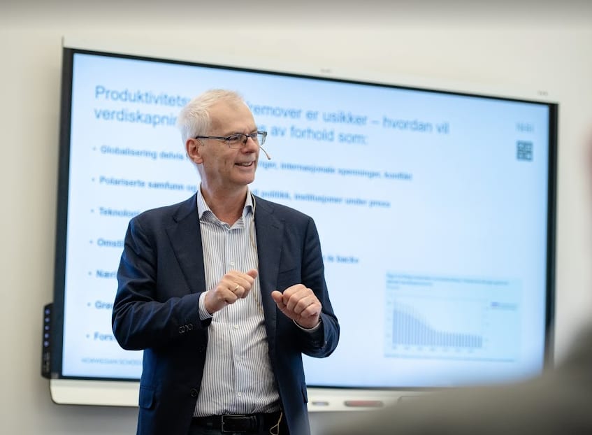 A smiling man in a suit and glasses presents in front of a large screen displaying presentation slides.