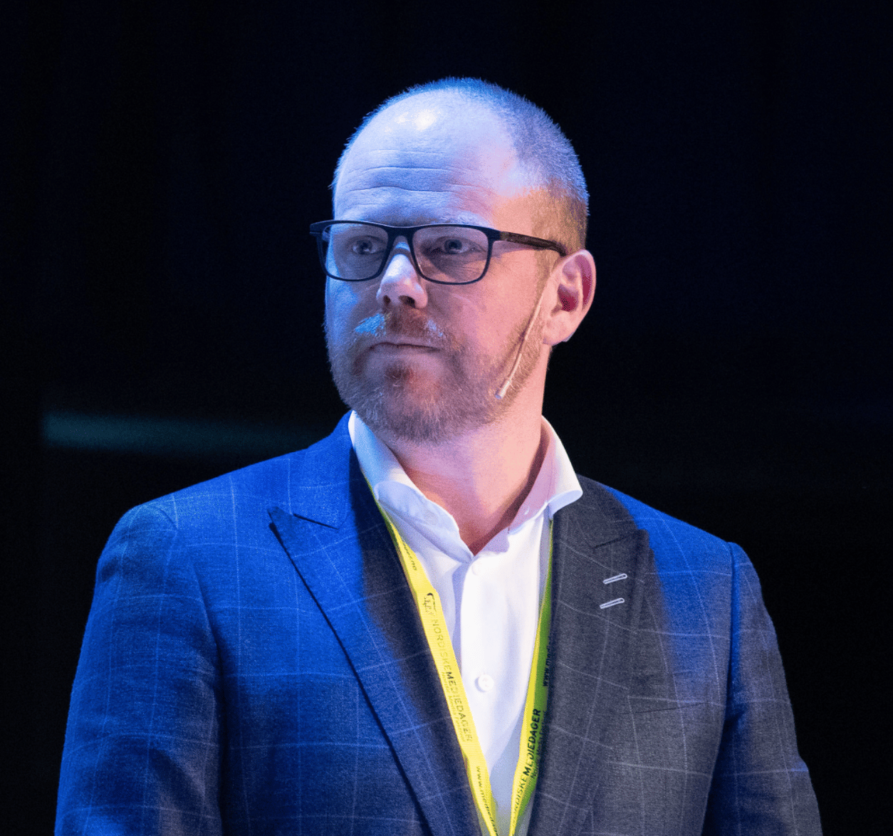 Bearded man in glasses, blue suit, with "Nordisk Mediedager" lanyard on stage.