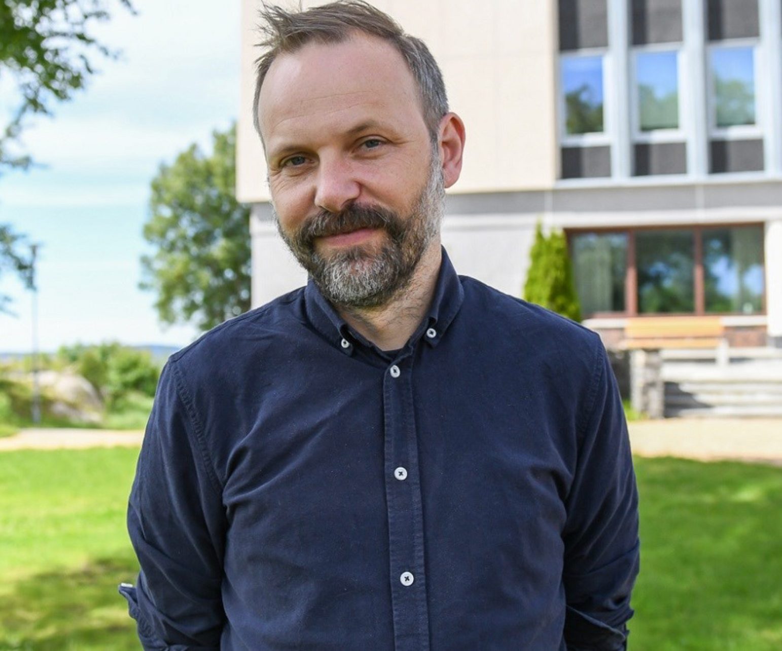 Smiling bearded man in blue shirt outdoors, with a building and trees behind.