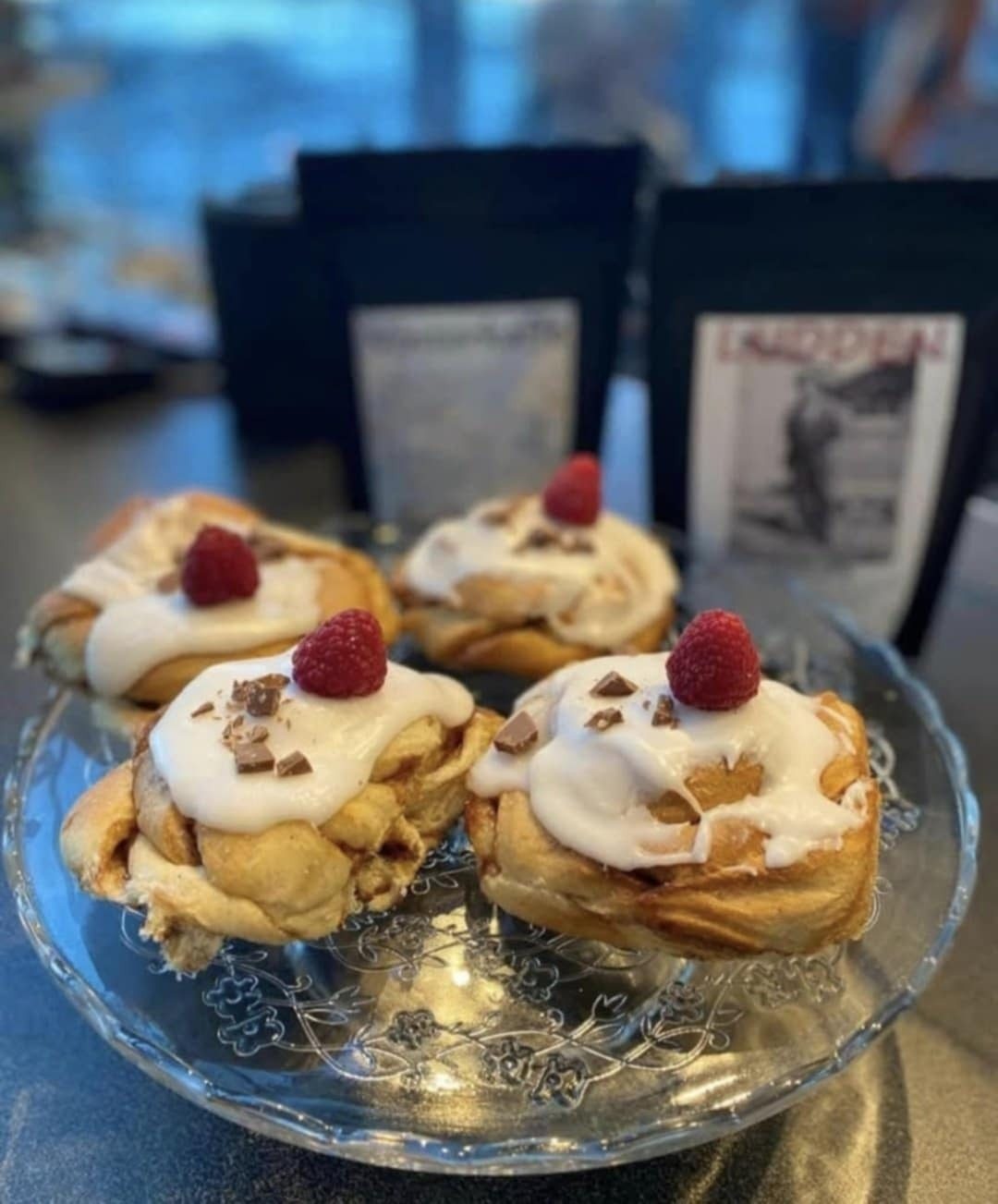 Four frosted cinnamon rolls with raspberries and chocolate on a glass plate.