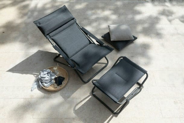 Dark gray outdoor lounge chair & footrest on a sunlit patio with tray and cushions.