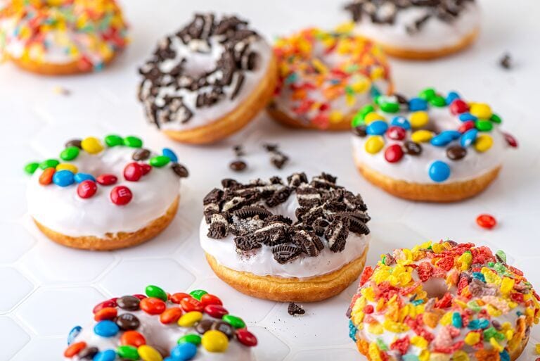 An assortment of colorful, glazed donuts topped with M&Ms, crushed Oreos, and various sprinkles on a white surface.