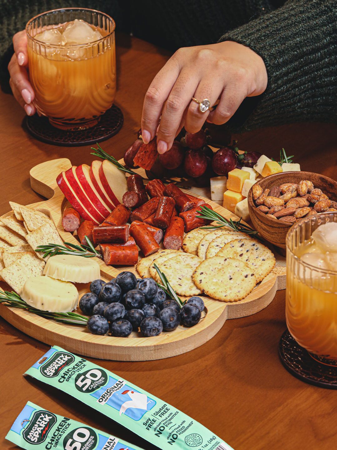 Hand picks snack from charcuterie board with meat sticks, cheese, fruit, crackers, drinks, and snack packs.