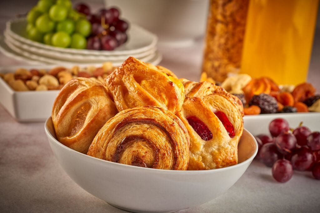 A close-up of a white bowl filled with assorted pastries, with grapes, nuts, and dried fruit in the background.