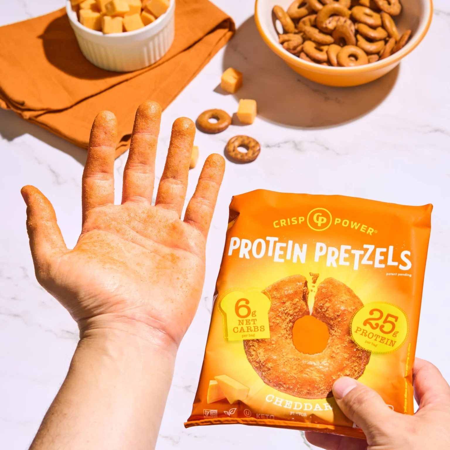 A hand covered in orange seasoning from cheddar protein pretzels, holding the snack bag next to bowls of pretzels and cheese.