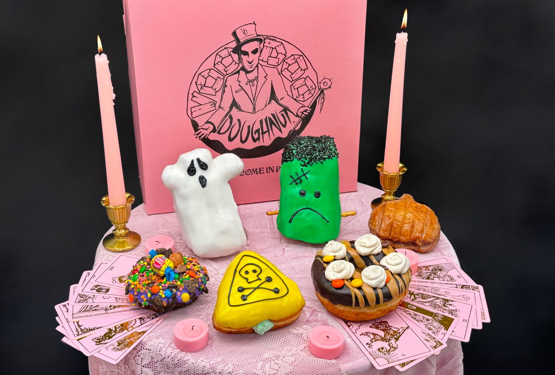 Halloween-themed donuts (ghost, Frankenstein, pumpkin, skull) on a pink tablecloth with candles and tarot cards.