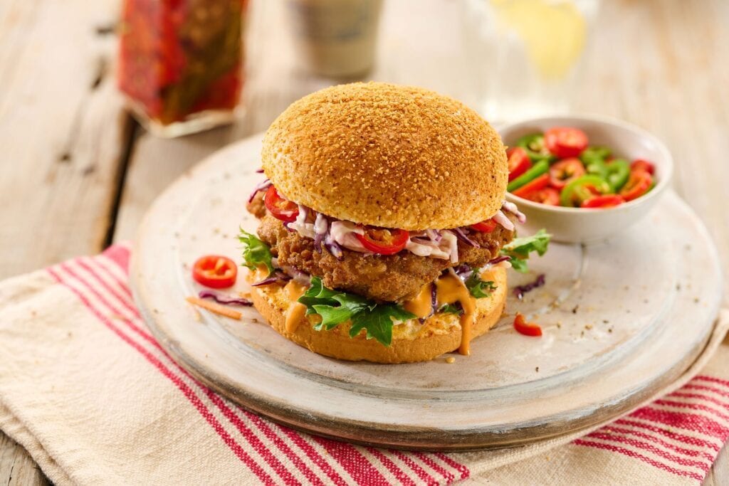 A crispy fried chicken sandwich topped with coleslaw, sliced peppers, and sauce, served on a bun.