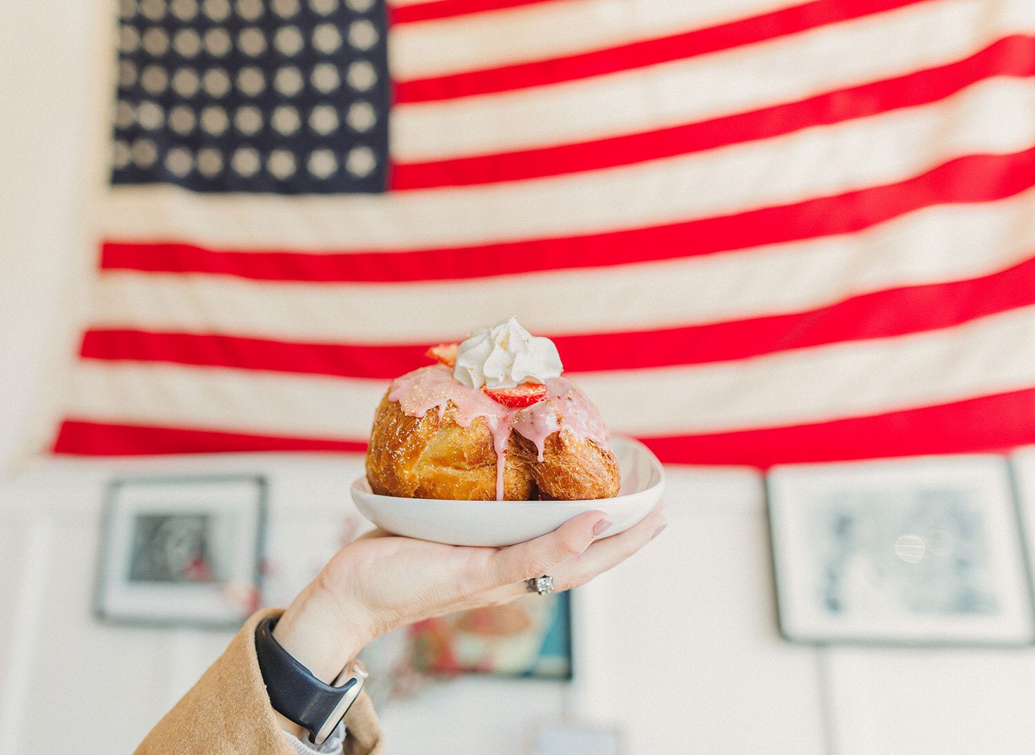 A hand holds a plate with a croissant covered in pink icing, whipped cream, and strawberries, against an American flag.