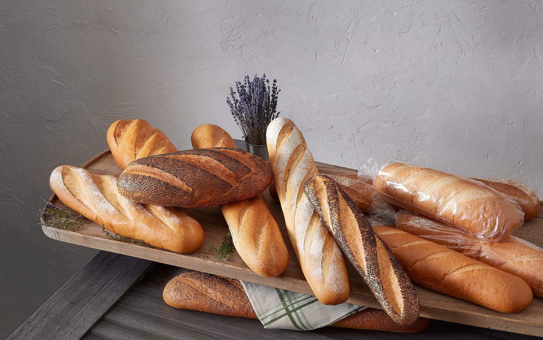 Assorted fresh bread loaves: plain, poppy seed, and wrapped, on a wooden tray with lavender.