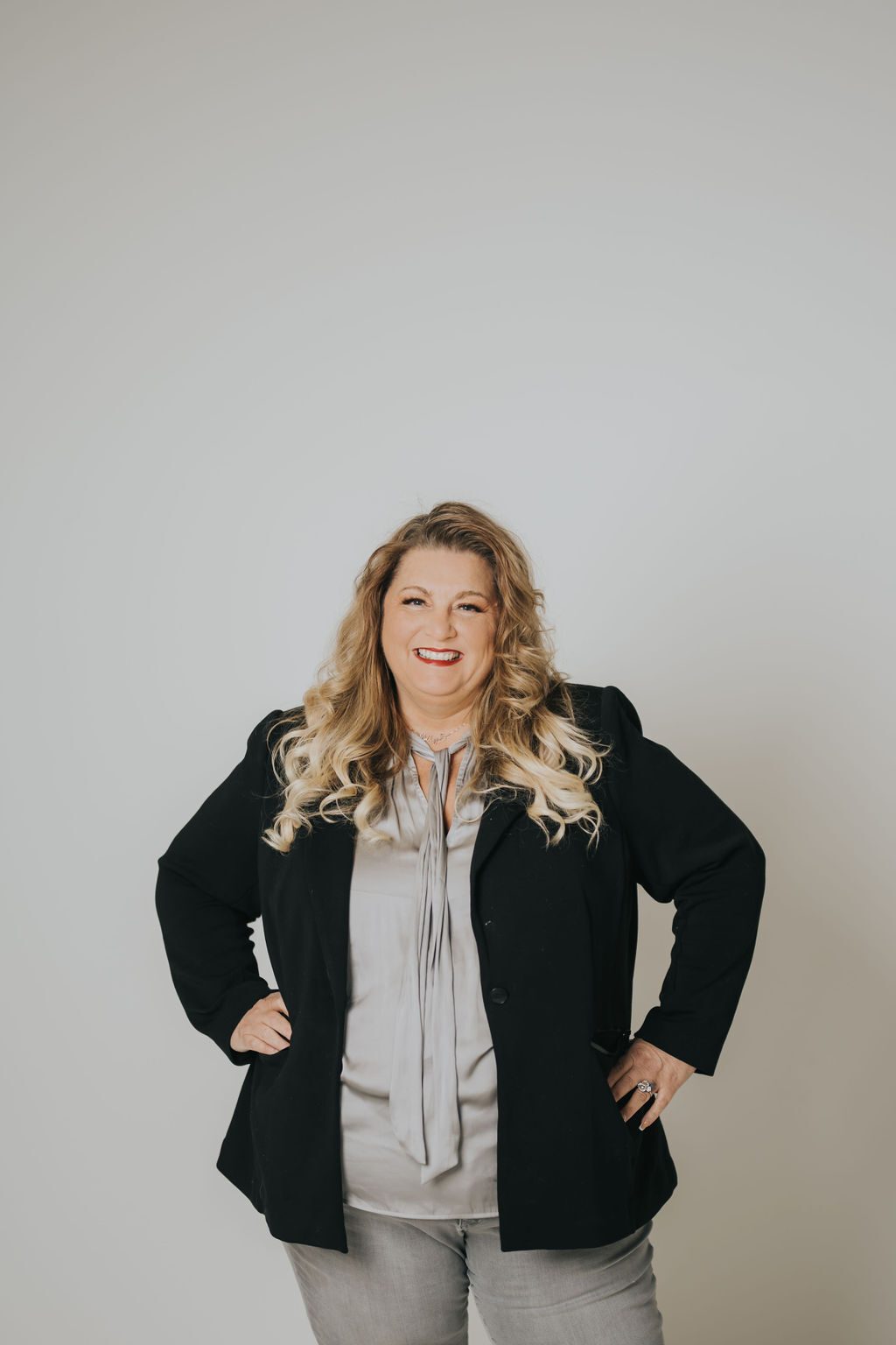 Smiling woman with long blonde curly hair in a black blazer and grey blouse, hands on hips.