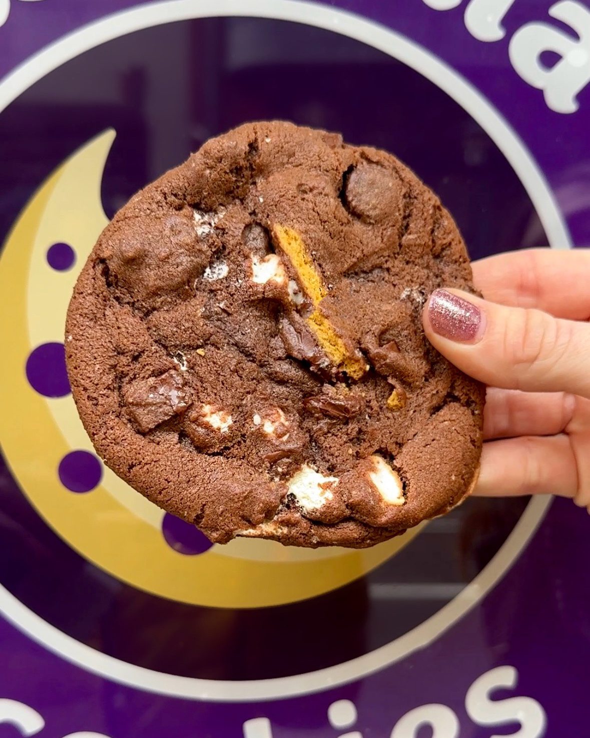 Hand holding a chocolate cookie with marshmallows and honeycomb against a purple background.