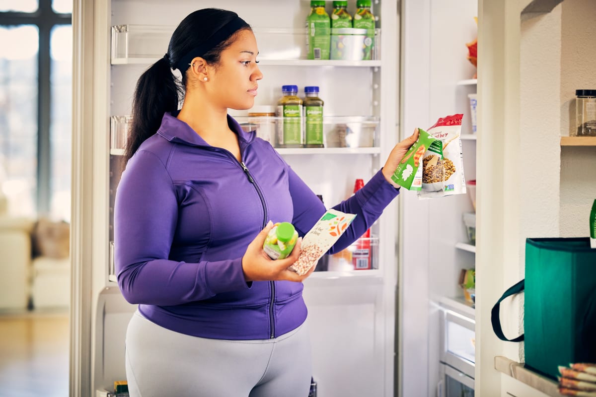 A woman wearing a purple tracksuit looks in her fridge at various healthy food items in packages