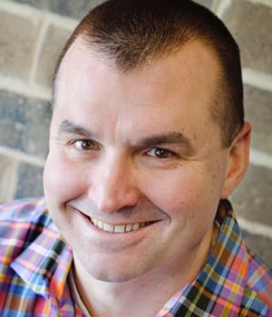 Smiling man with short hair and a receding hairline wearing a colorful plaid shirt, looking at the camera.