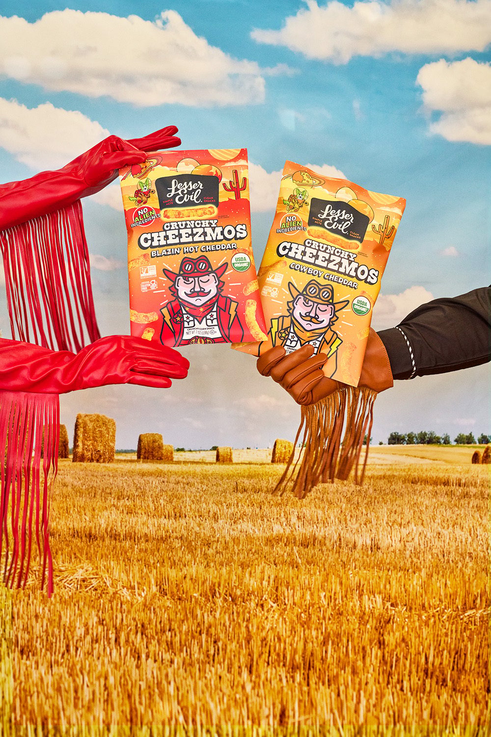 Two fringed-gloved hands hold LesserEvil Crunchy Cheezmos bags in a golden field with hay bales under a blue sky.