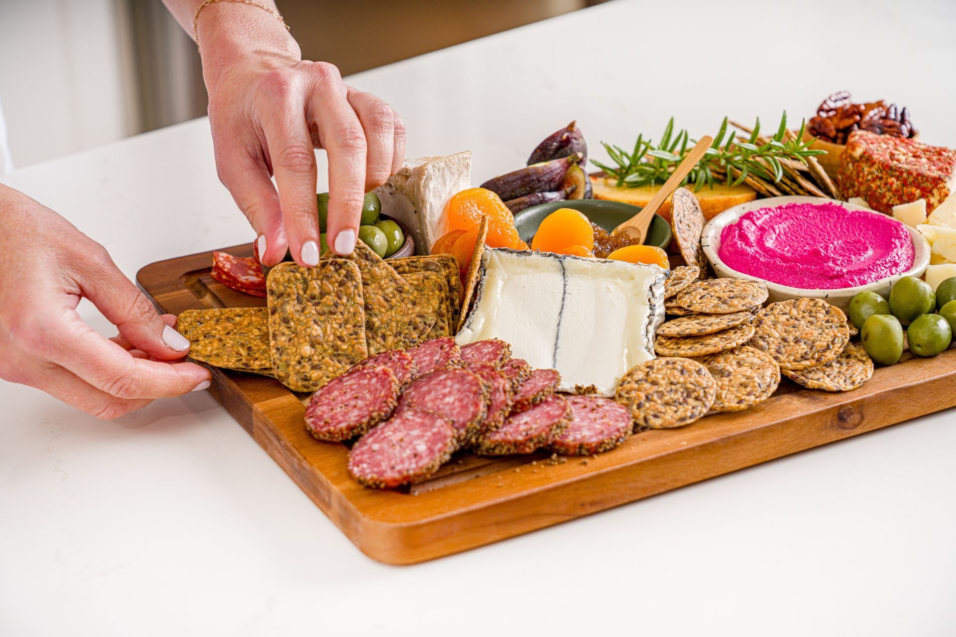 Hands arrange crackers on a vibrant charcuterie board with cheese, salami, olives, fruits, and dip.