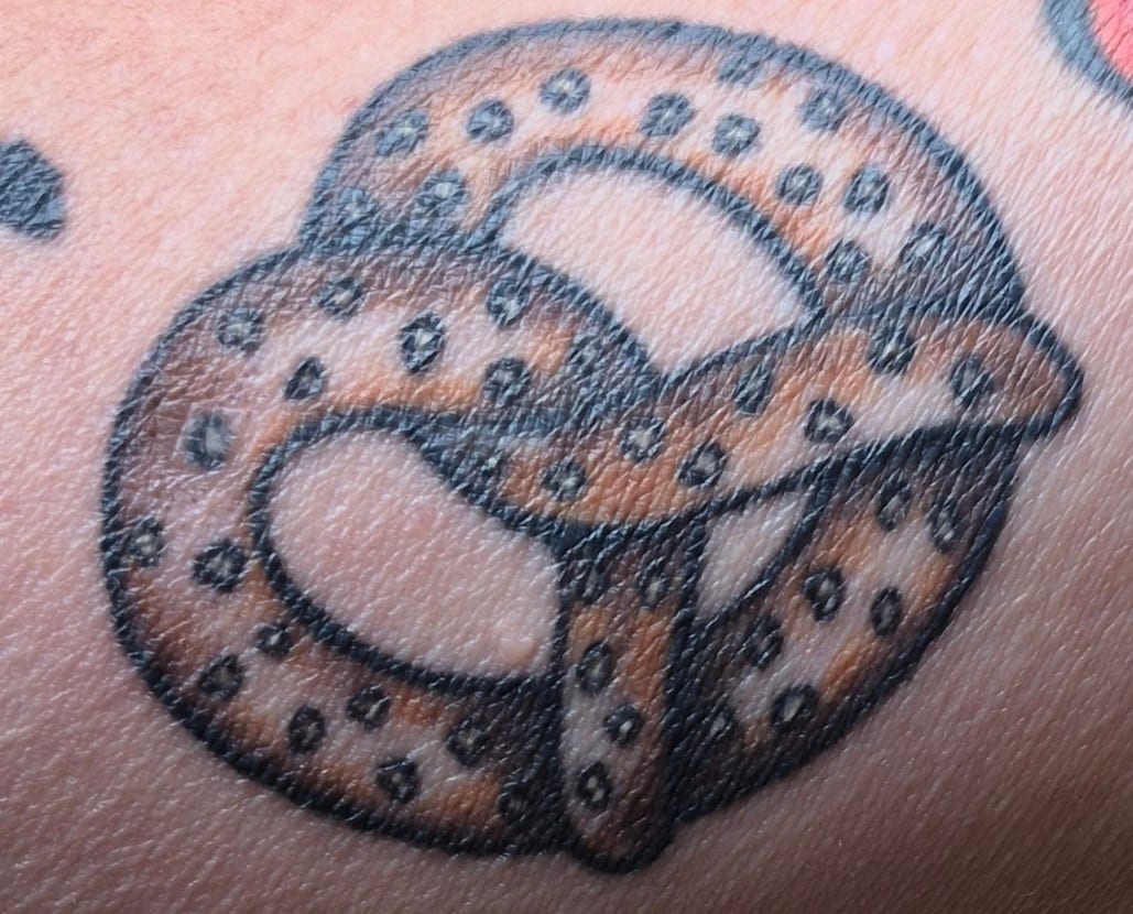 A brown pretzel tattoo with black outlines and salt dots on skin.