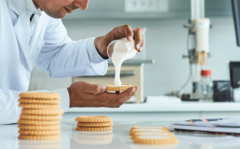 Lab professional in white coat applies cream to a cookie; more cookies, lab equipment.