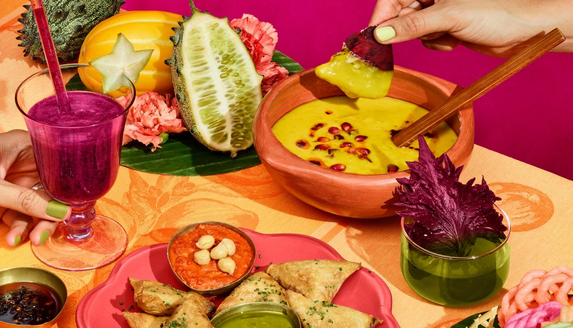 Colorful feast: hand dipping chip into yellow dip, purple smoothie, samosas, and exotic fruits.