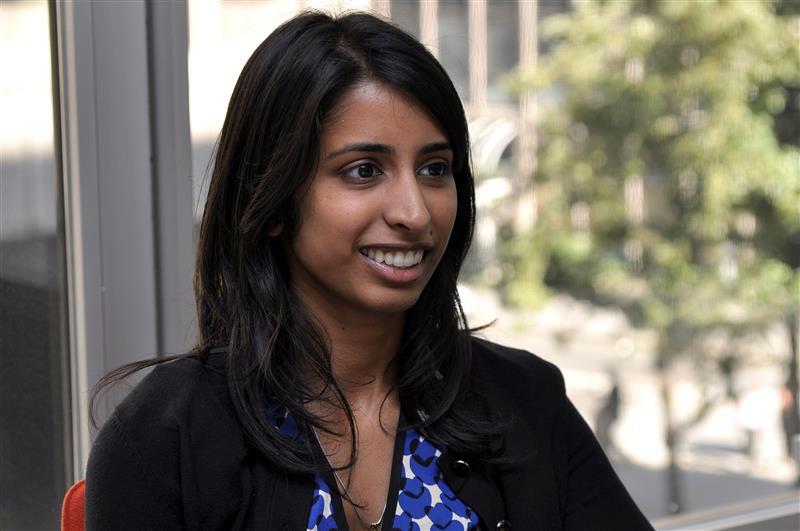 Smiling woman with dark hair in black cardigan, looking right, with blurred city background.