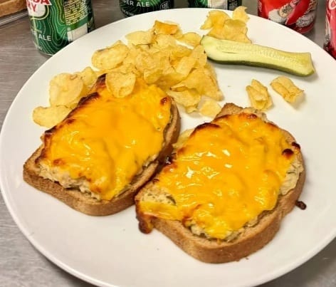 Two open-faced tuna melts on toast with melted cheese, served with potato chips and a pickle spear.