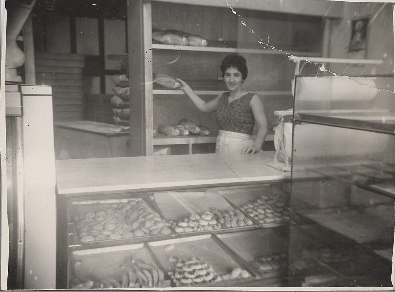 Smiling woman in a bakery holding bread, with pastries visible in a display case.