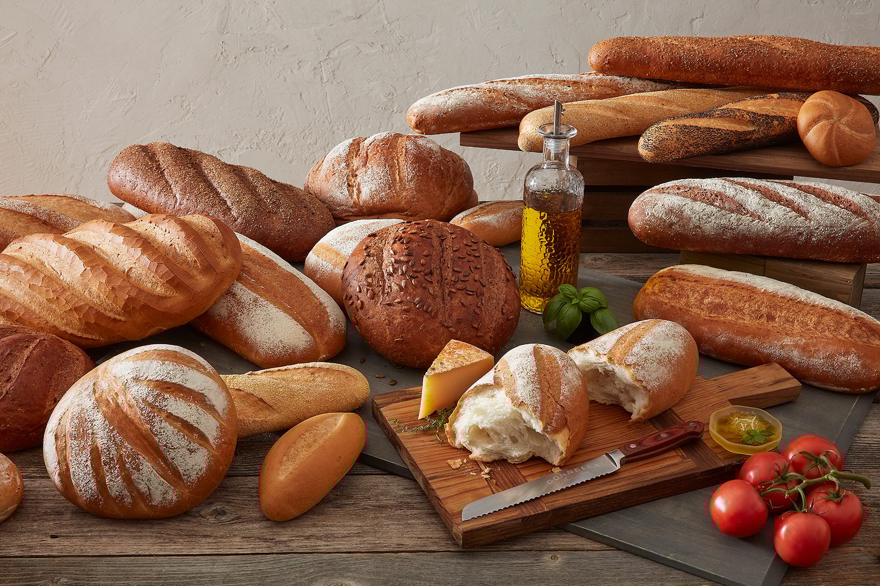 Variety of fresh breads, olive oil, basil, tomatoes, and cheese on a wooden table.