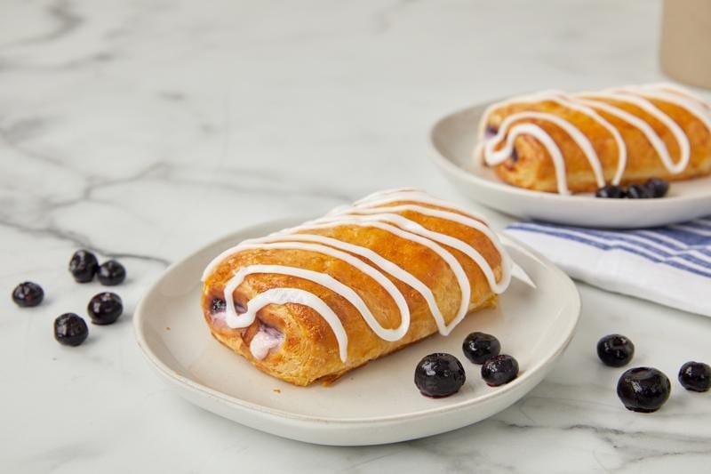 Two iced blueberry danishes on white plates with fresh blueberries.