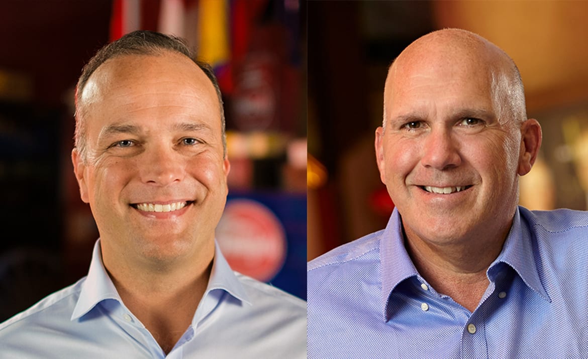 Two smiling men in blue shirts: one with dark hair, one bald.