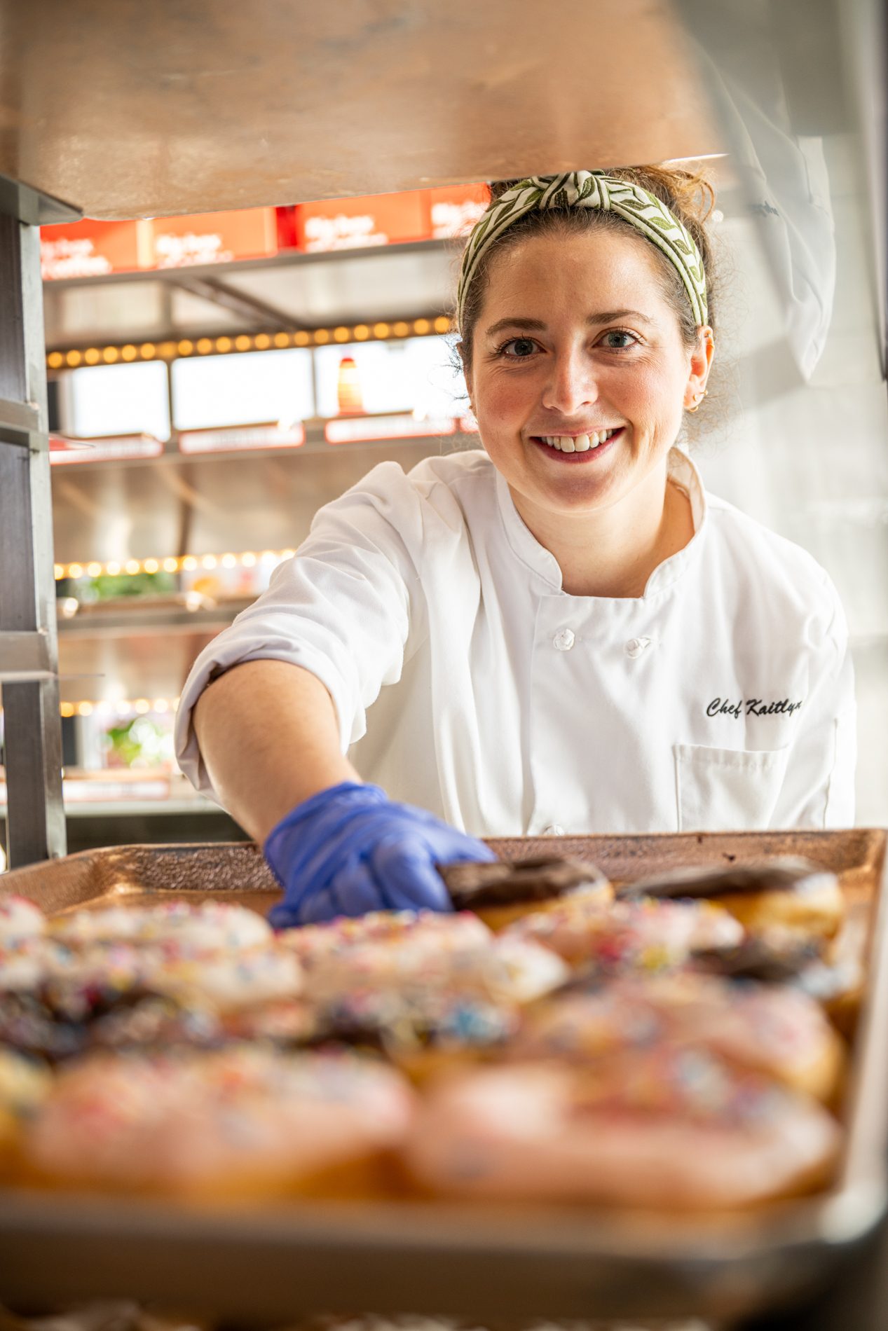 Smiling chef in white coat and blue glove arranging sprinkle donuts on a tray.