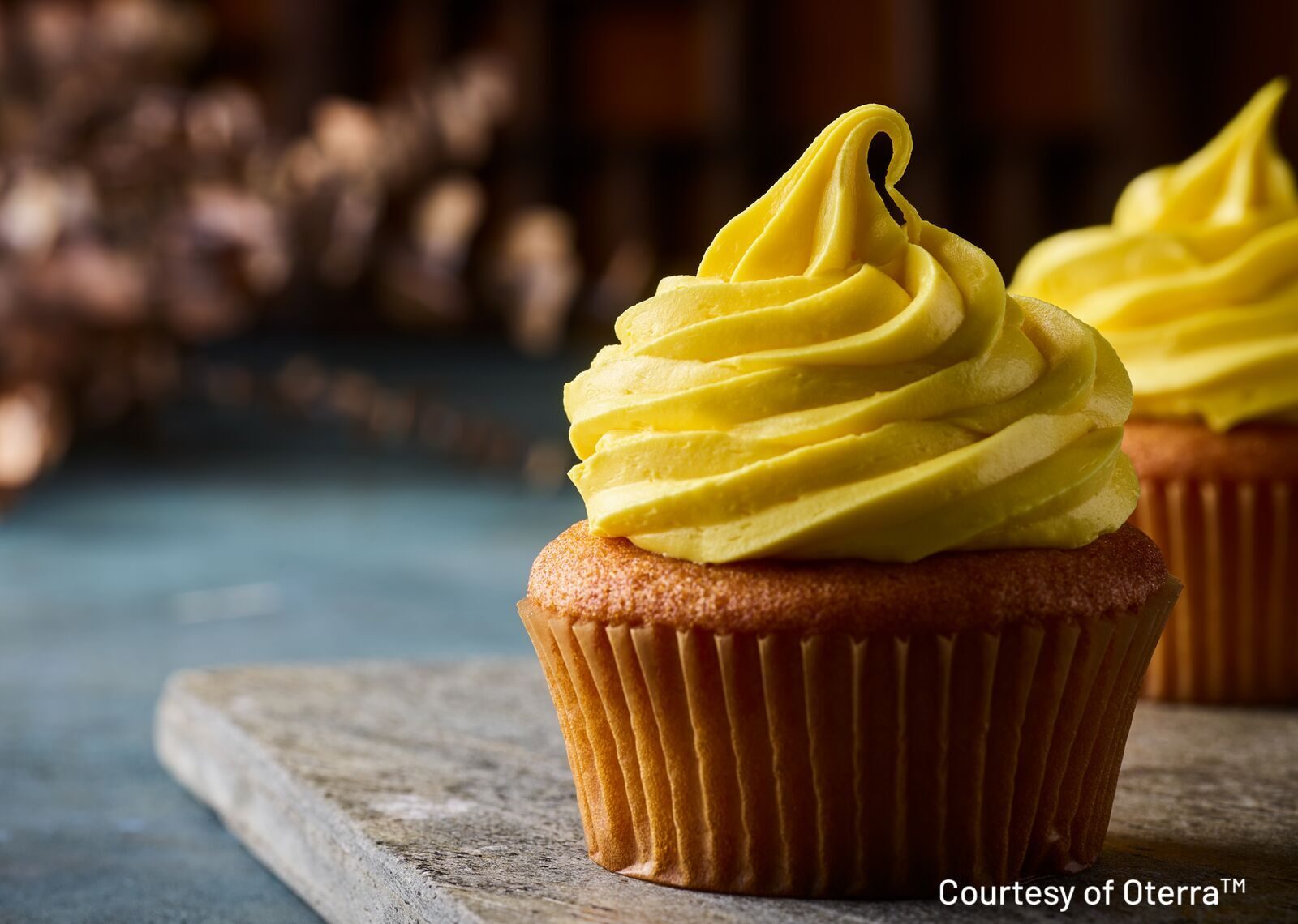 Two freshly baked cupcakes with bright yellow frosting, one in focus and another blurred behind.