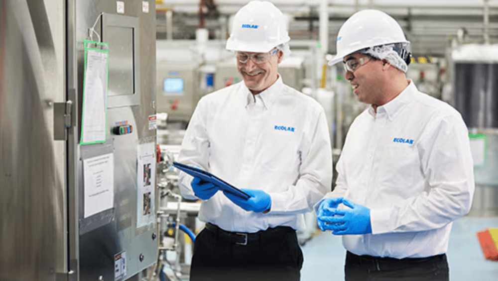 Two Ecolab employees in hard hats and gloves reviewing a tablet in a plant.
