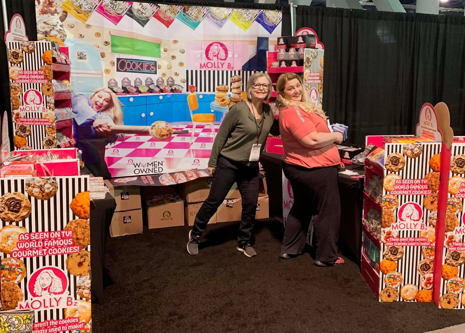 Two women pose at a vibrant "Molly B's World Famous Gourmet Cookies" trade show booth.